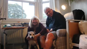 A smiling couple sits indoors by a window, with a small dog in the woman's arms. The room is cozy, with soft light creating a warm atmosphere.