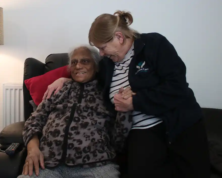 An elderly woman in a leopard-print sweater sits smiling on a couch, holding hands with a younger woman. Both share a warm, affectionate moment.